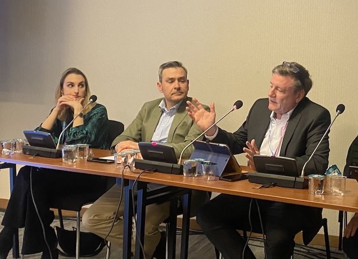 Man jesticulates behind table as two other sitting by him look on