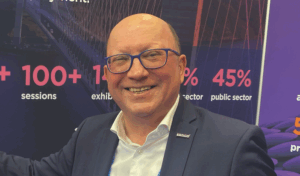 Bald white man in classes smiles broadly against a backdrop of some stats. He wears and open neck shirt and had a suit jacket with an ERTICO pin page