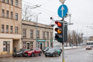 A traffic light displaying red and amber signals at an urban junction in Riga, Latvia, with surveillance and monitoring cameras mounted on the pole alongside a blue directional road sign. Several parked cars and city buildings are visible in the background on an overcast winter day.