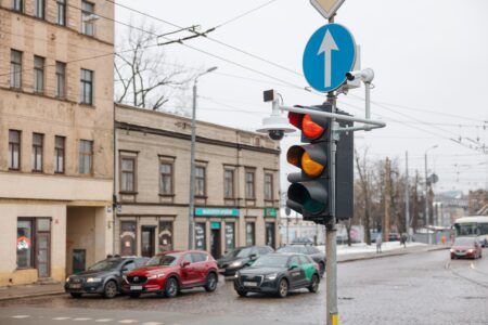 A traffic light displaying red and amber signals at an urban junction in Riga, Latvia, with surveillance and monitoring cameras mounted on the pole alongside a blue directional road sign. Several parked cars and city buildings are visible in the background on an overcast winter day.