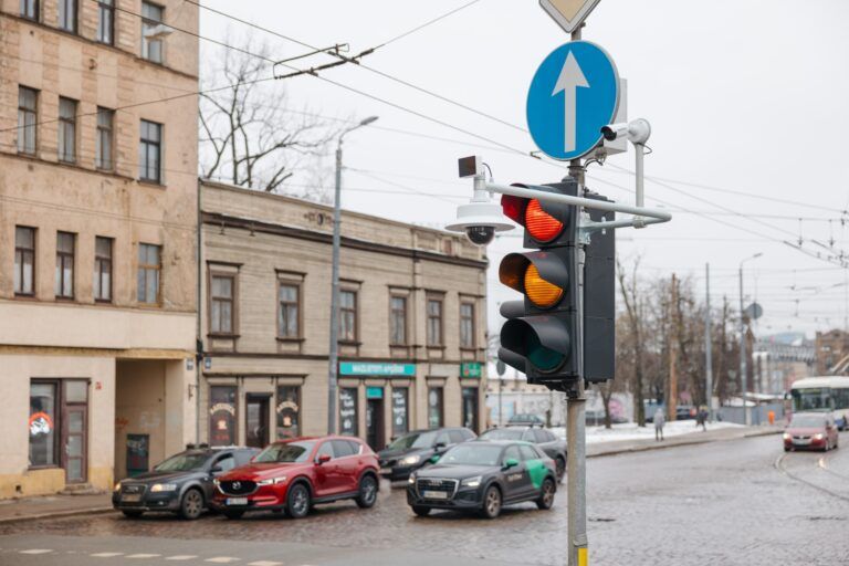 A traffic light displaying red and amber signals at an urban junction in Riga, Latvia, with surveillance and monitoring cameras mounted on the pole alongside a blue directional road sign. Several parked cars and city buildings are visible in the background on an overcast winter day.