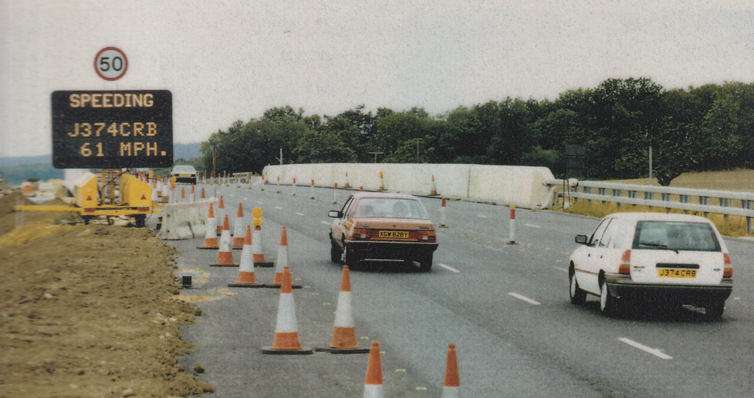 Motorway roadworks scene with orange traffic cones and concrete barriers, showing a variable message sign displaying "SPEEDING J374CRB 61 MPH" beneath a 50mph speed limit sign, with two cars passing through the works