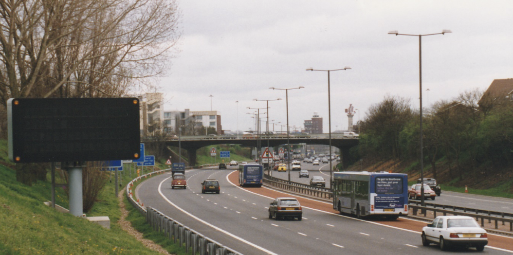 Wide view of the M4 Spur road approaching Heathrow Airport showing a dedicated bus lane marked in red, a large variable message sign on the left verge, a bus and several cars on the carriageway, with the Heathrow control tower visible in the background