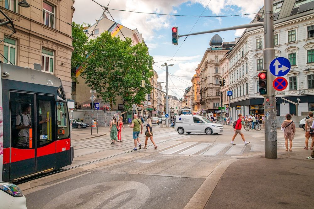 Busy city intersection with a tram, pedestrians crossing, cyclists, a van and traffic signals showing red.