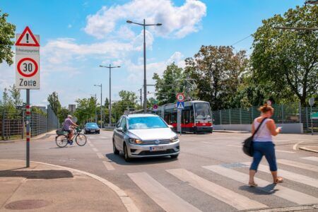 A 30km/h zone sign next to a road junction with a car, a tram, a cyclist and a pedestrian crossing on a zebra crossing.