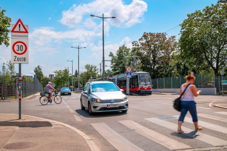 A 30km/h zone sign next to a road junction with a car, a tram, a cyclist and a pedestrian crossing on a zebra crossing.