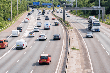 Busy UK motorway with multiple lanes of traffic including cars, vans and a car transporter, with overhead gantry signs for the M61, M62, M65 and M58.