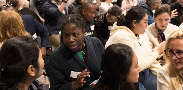 Attendees engaged in animated discussion at a round-table event, with a woman in a dark blazer gesturing as she makes a point to fellow delegates.