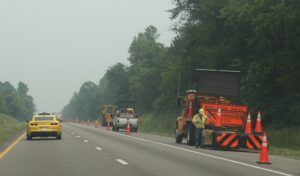 A road worker in high-visibility clothing operates equipment at the side of a multi-lane highway, with orange traffic cones, a "Road Work Ahead" sign, and a shoulder closure board visible. A yellow car passes in the adjacent lane.