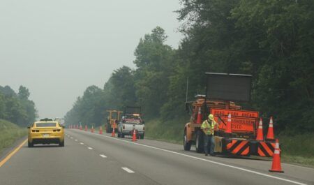 A road worker in high-visibility clothing operates equipment at the side of a multi-lane highway, with orange traffic cones, a "Road Work Ahead" sign, and a shoulder closure board visible. A yellow car passes in the adjacent lane.