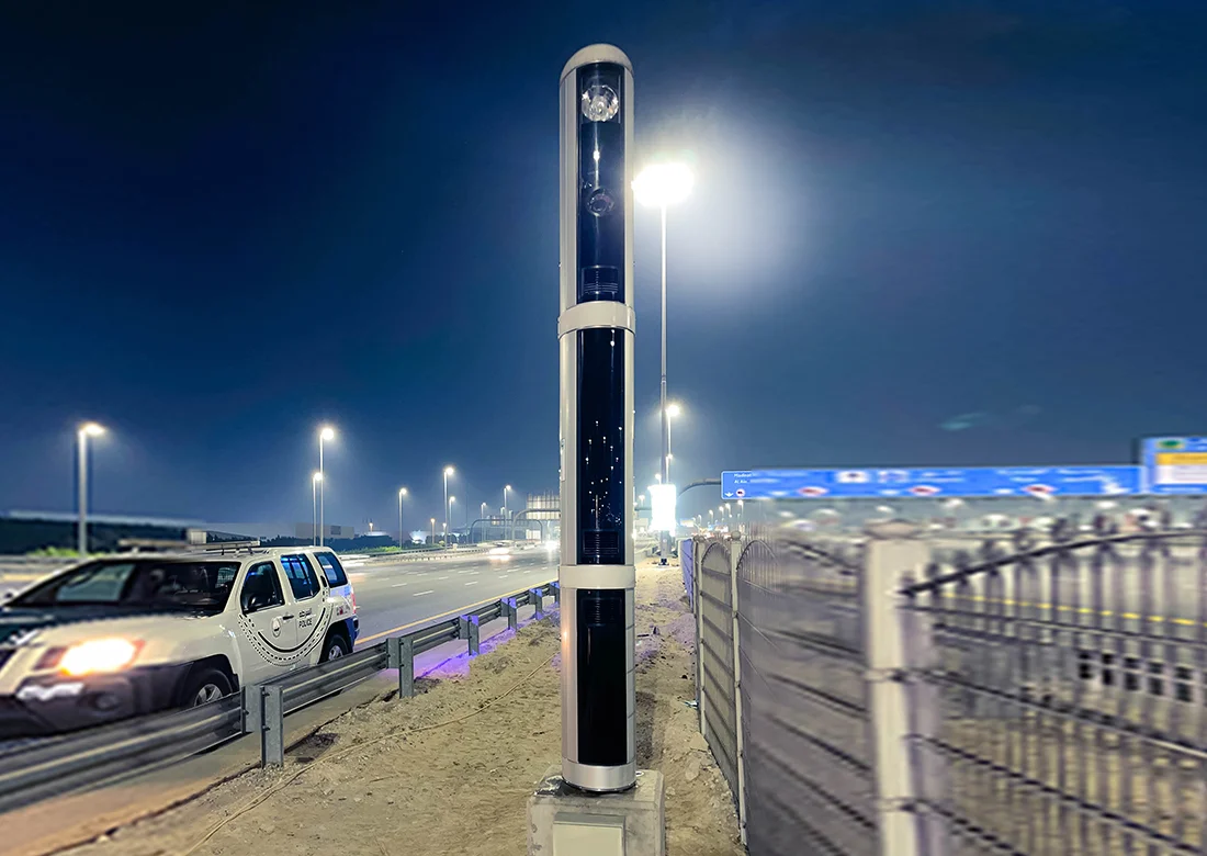 A tall cylindrical roadside enforcement unit installed beside a multi-lane highway at night, with a police vehicle and airport signage visible in the background.