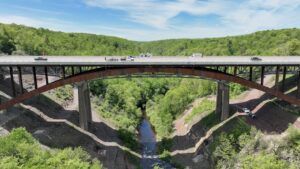 Aerial side view of a highway bridge carrying vehicles including a flatbed truck, with orange traffic cones along one carriageway and construction activity visible at the far end