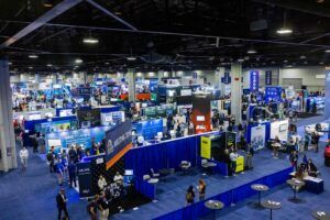 Elevated wide-angle view of a large, busy exhibition hall with numerous exhibitor stands, banners and attendees visible across the floor.