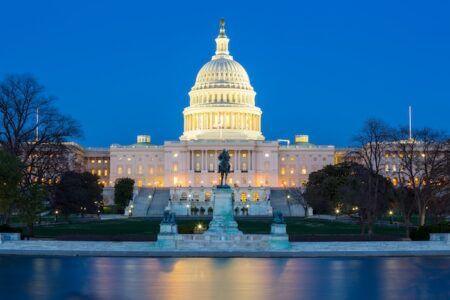 The United States Capitol building illuminated at dusk, reflected in the Capitol Reflecting Pool, with the dome lit against a deep blue sky.