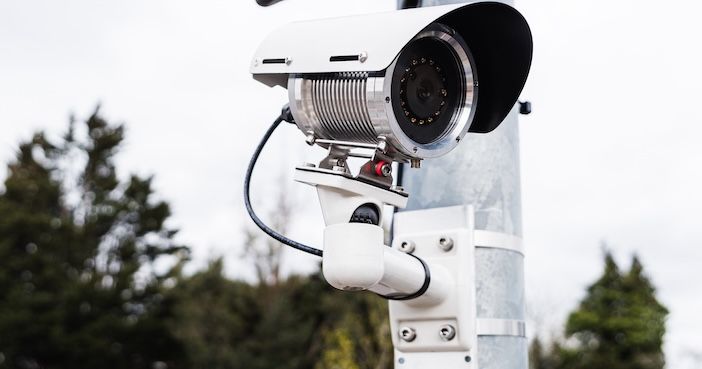 A bullet-style ANPR camera mounted on a galvanised metal pole at a car park entrance, with parked vehicles visible in the blurred background and trees beyond.