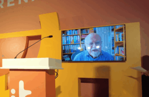 A screen mounted on an orange and yellow stage wall displays a video call of an elderly man with a white beard and glasses wearing a blue striped shirt, seated in front of a bookcase filled with coloured folders and books. A microphone and the edge of a white podium are visible to the left of the screen.