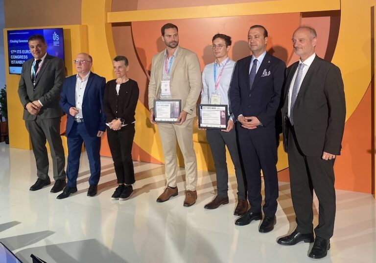 : Seven people stand in a line on a stage in front of an orange and yellow curved backdrop. A screen on the left displays '17th ITS European Congress Istanbul — Closing Ceremony'. Two of the people in the centre hold framed award certificates. The group includes men in suits and one woman in dark clothing