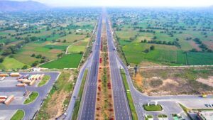 Aerial view of a wide, multi-lane divided highway in India, stretching to the horizon through a flat agricultural landscape, with a large lorry parking area visible to the left.