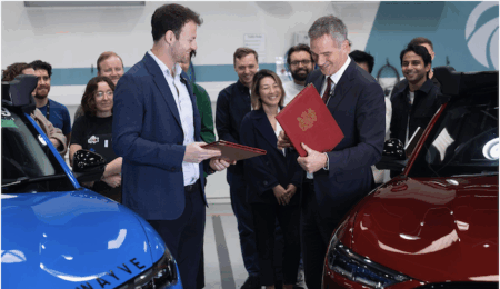 Two men exchange red-covered document folders in an indoor setting surrounded by a group of onlookers, with two vehicles — one blue, one red — visible in the foreground
