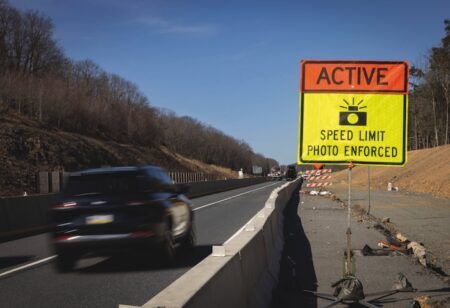 An 'ACTIVE speed limit photo enforced' sign in a road work zone, with a car passing with speed blur in the photo