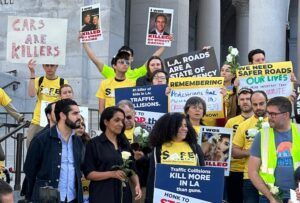 Protesters gather on steps outside a public building holding placards calling for safer roads in Los Angeles. Signs include messages such as "Cars are killers", "Traffic collisions kill more in LA than guns" and "LA roads are a state of emergency". Several demonstrators wear yellow T-shirts bearing the Streets Are For Everyone (Safe) logo. Some hold white roses and photographs of people killed in traffic collisions.