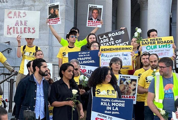 Protesters gather on steps outside a public building holding placards calling for safer roads in Los Angeles. Signs include messages such as "Cars are killers", "Traffic collisions kill more in LA than guns" and "LA roads are a state of emergency". Several demonstrators wear yellow T-shirts bearing the Streets Are For Everyone (Safe) logo. Some hold white roses and photographs of people killed in traffic collisions.