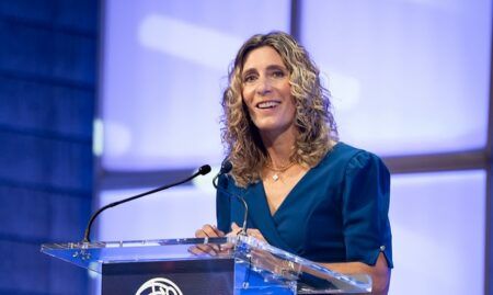 A woman with long blonde curely hair speaking at a podium on a conference stage, smiling, wearing a teal dress
