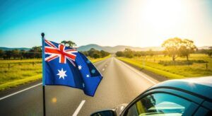 An Australian flag mounted on a car window, with a straight rural road stretching into the distance through open green fields, trees and hills under a bright blue sky
