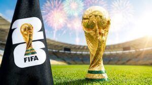 A black FIFA World Cup 2026 corner flag with the number 26 stands alongside the gold FIFA World Cup trophy, with stadium fireworks in the background