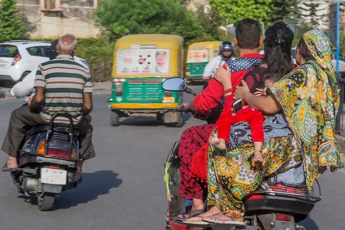 Rear view of four people including a young child riding together on a single motorcycle in Indian city traffic, with auto-rickshaws and other vehicles visible ahead.