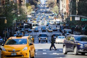 An elevated view looking north along a busy multi-lane New York City avenue, with yellow taxis, SUVs, a bus and other vehicles filling the carriageway. A pedestrian crosses in the foreground. Cross-street signs for E 47th and E 49th Streets are visible, along with traffic signals and tree-lined pavements.