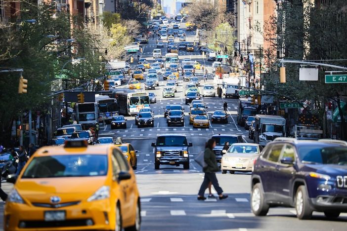 An elevated view looking north along a busy multi-lane New York City avenue, with yellow taxis, SUVs, a bus and other vehicles filling the carriageway. A pedestrian crosses in the foreground. Cross-street signs for E 47th and E 49th Streets are visible, along with traffic signals and tree-lined pavements.