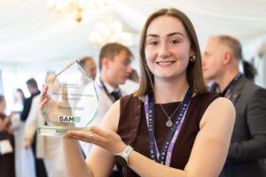 A young woman with brown shoulder-length hair smiles at the camera while holding a glass flame-shaped award engraved with "Intelligent Transport Systems, Allan Prize National Essay Competition, Winner 2026" and the SAM logo. She is wearing a Mobius Networks visitor lanyard.