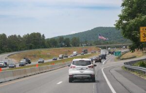 ehicles travelling on a multi-lane US interstate highway in a rural setting, with orange construction barriers visible along the central reservation, an American flag in the background, and a green exit sign for Exit 146 on the right.