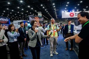 A woman holding a microphone speaks animatedly to a crowd of attendees on the exhibition floor at ITS America 2026, with the Taiwan Excellence stand and Trafficvision banner visible in the background. Several delegates look on, some smiling.