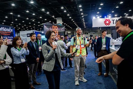 A woman holding a microphone speaks animatedly to a crowd of attendees on the exhibition floor at ITS America 2026, with the Taiwan Excellence stand and Trafficvision banner visible in the background. Several delegates look on, some smiling.