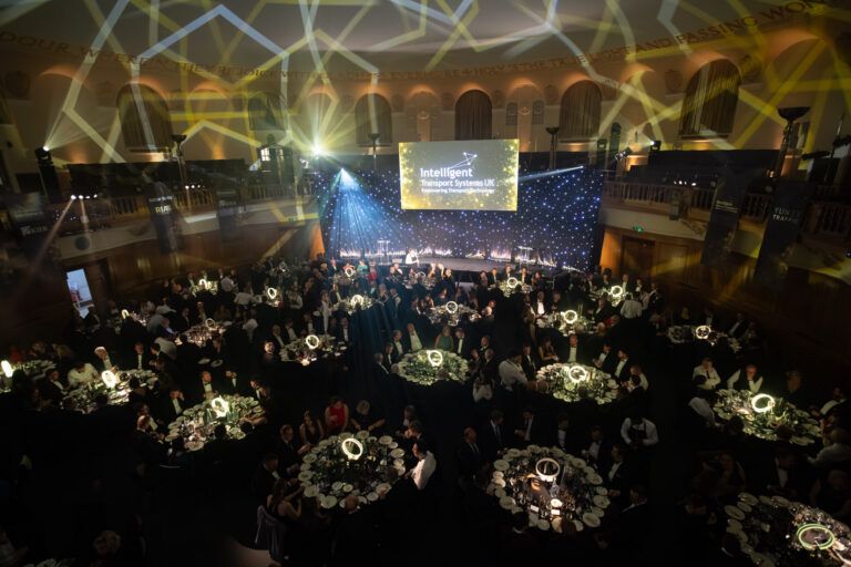 Aerial view of a large formal dinner in a grand hall, with round tables of guests in black tie, stage lighting and a large screen displaying the Intelligent Transport Systems UK logo at the front
