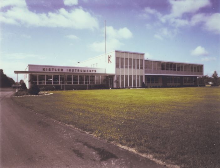 Archive colour photograph of a single-storey mid-century building with a "Kistler Instruments" sign and flagpole, set behind an open lawn