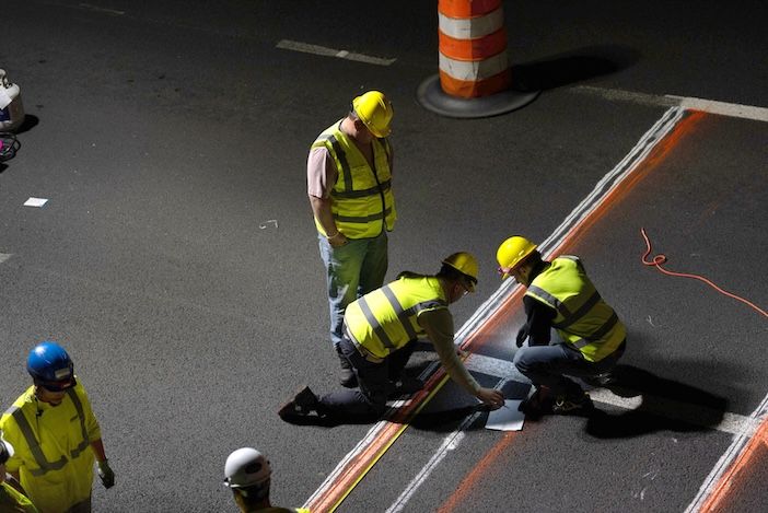 Overhead night-time view of a road crew in yellow hi-vis vests and hard hats kneeling to install sensor equipment into a road surface marked with orange guide lines, next to a traffic cone