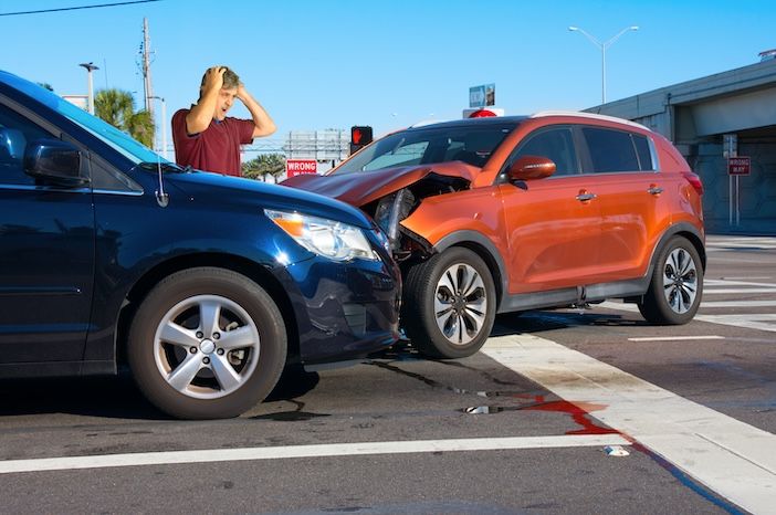 Two cars have crashed wrecked into each other at intersection with very upset man driver looking at the severe damage with wrong way sign in background.