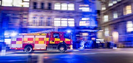 A red and yellow chequered fire and rescue service engine drives at speed through a city street at night, with motion blur streaking the illuminated building facades and blue emergency lights behind it.