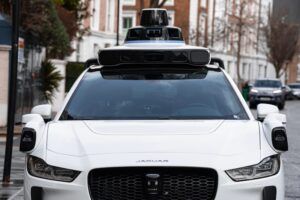 A white Jaguar I-PACE autonomous test vehicle fitted with roof-mounted sensor and camera equipment, parked on a residential London street, viewed from the fron