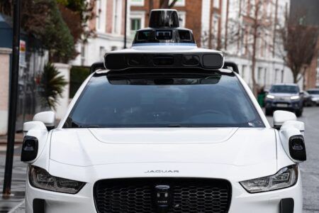 A white Jaguar I-PACE autonomous test vehicle fitted with roof-mounted sensor and camera equipment, parked on a residential London street, viewed from the fron