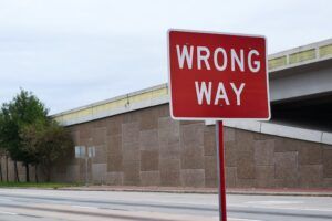 A red and white "Wrong Way" road sign mounted on a pole beside a road, with an overpass in the background