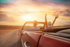 A man and woman drive a red convertible along a dirt road at sunset, with the woman raising both arms in the air
