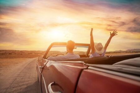 FEATURE: Drivers trust road safety tech more than experts, study finds A man and woman drive a red convertible along a dirt road at sunset, with the woman raising both arms in the air