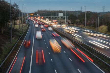 National Highways meets targets but RIS3 risks flagged Long-exposure night view of a UK motorway showing red brake-light trails in the near carriageway and white headlight trails in the opposite carriageway, with an overhead electronic gantry sign and street lighting either side