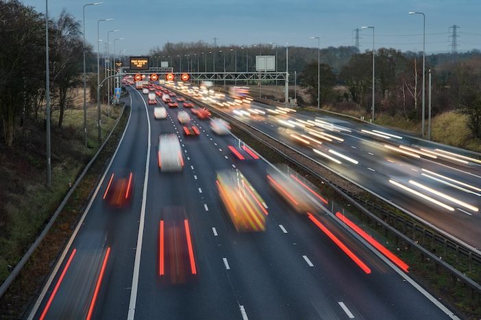 Long-exposure night view of a UK motorway showing red brake-light trails in the near carriageway and white headlight trails in the opposite carriageway, with an overhead electronic gantry sign and street lighting either side