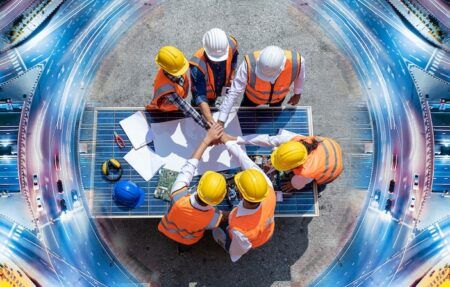 Six workers in orange high-visibility vests and yellow, white and blue hard hats join hands above a solar panel on a worktop surrounded by engineering plans. The background is a digitally composited aerial view of illuminated motorway interchanges.