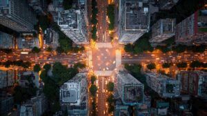 An aerial night-time view looking straight down on a busy city road intersection, with light trails from traffic and illuminated high-rise buildings on each corner